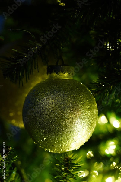 Fototapeta Glittery golden ball on artificial Christmas tree. Vertical greeting card template. Shiny bauble hanging on a branch of plastic Xmas tree in the dark, lit by garlands. Close-up, blurred background.