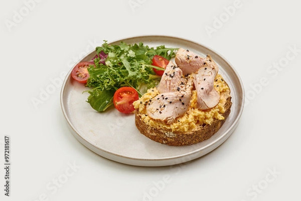 Fototapeta Breakfast plate featuring a slice of whole-grain toast topped with scrambled eggs, smoked turkey, and sesame seeds. A fresh side salad of cherry tomatoes and greens adds a vibrant touch.
