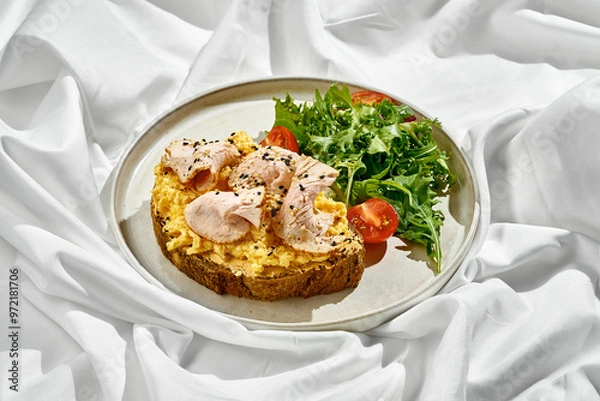 Fototapeta Breakfast plate featuring a slice of whole-grain toast topped with scrambled eggs, smoked turkey, and sesame seeds. A fresh side salad of cherry tomatoes and greens adds a vibrant touch.
