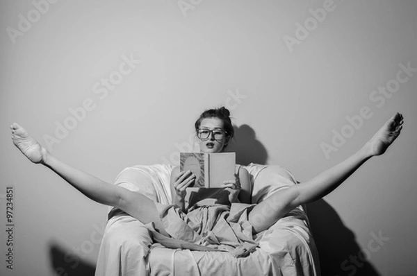 Fototapeta Black and white photography of beautiful young lady wearing glasses and holding a book relaxing split legs in chair