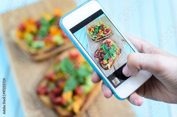 Fototapeta Taking photo on smartphone of fresh homemade rustic bruschetta with chopped tomatoes, basil and olive oil on toasted garlic bread on wooden board over turquoise background