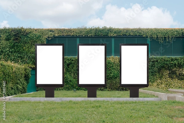 Fototapeta Vertical mock up template: 3 out of home (OOH) advertising billboard posters on grass, against background of lush plant wall of building. Bright day, blue sky with clouds. Transparent png file.