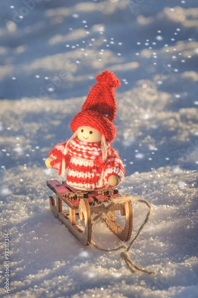 Fototapeta Little doll toy in knitted sweater and hat on toy wooden sleigh in evening light on snow. Christmas card. Vertical shot with copy space
