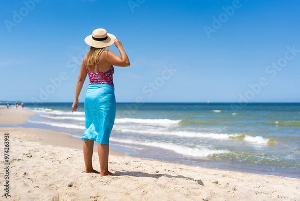 Obraz Summer vacation. Tanned woman in colorful swimsuit, blue pareo and sun hat standing by shore on sandy beach on beautiful sunny day. Back view	