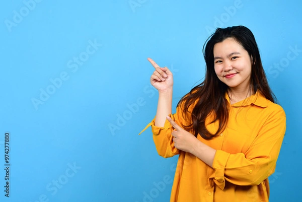 Fototapeta Happy asian woman in orange shirt pointing right side, left side and up side to copy space and looking at the camera isolated blue background