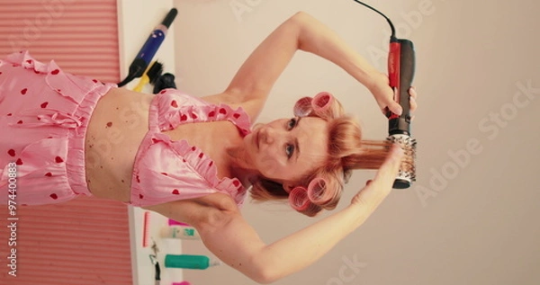 Fototapeta The girl makes curls with an electric curling iron. Warm styling of hair with a curling iron, independently. Vertical shot of a girl in pink on a pink background with an electric curling iron.
