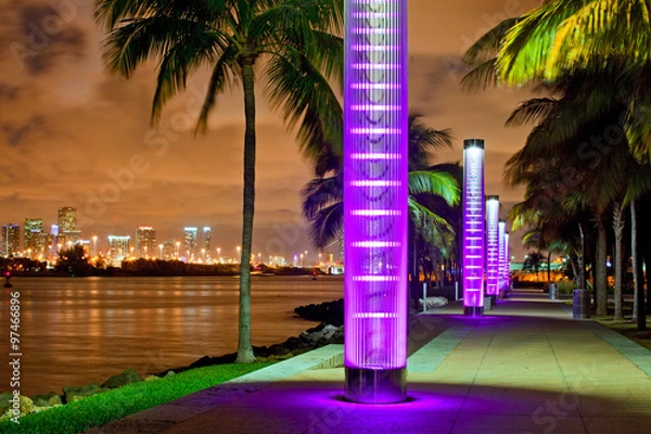 Fototapeta Miami BEach Florida at night, South Pointe park colorful lights and pedestrian path by the ocean and panorama of downtown in the background. Famous tourist destination.