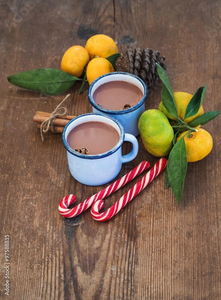 Fototapeta Hot chocolate in enamel metal mugs, fresh mandarins, cinnamon sticks, pine cone and candy canes over rustic wooden background