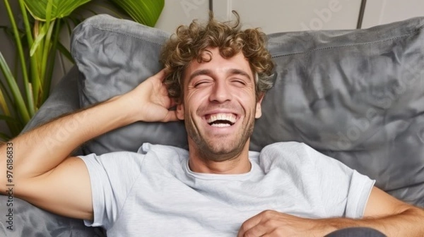 Obraz A young man with curly hair smiles widely while lying back on a comfortable couch. Sunlight filters through, highlighting a relaxing indoor atmosphere filled with greenery