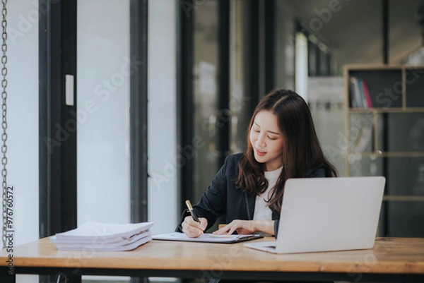 Fototapeta A cheerful young Asian businesswoman sits at her modern desk, typing on a laptop, working on corporate projects with paperwork, smiling in a busy workplace.