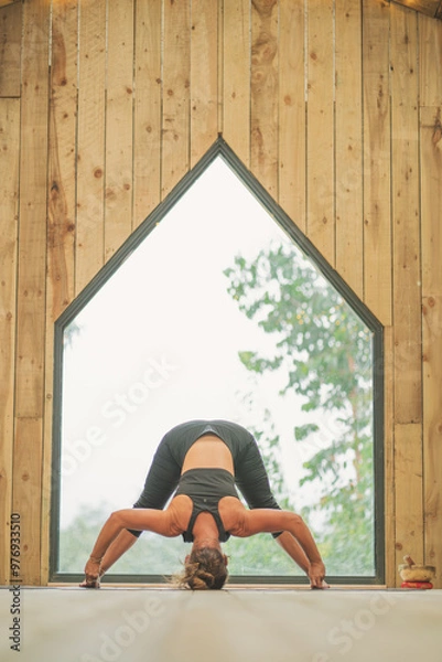 Obraz Woman doing Vinyasa yoga pose close to a window with black fitness outfit wooden room natural light wide leg forward pose D
