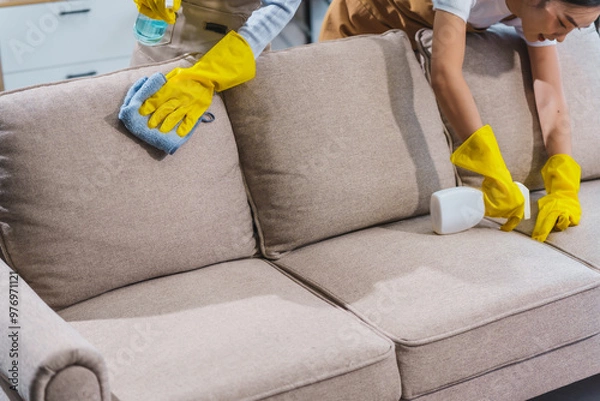 Fototapeta Two Asian housekeepers, wearing aprons and rubber gloves, happily clean an office. They sweep the floor, wipe glass surfaces, tidy up the sofa using cleaning liquids, a broom, and microfiber cloths.