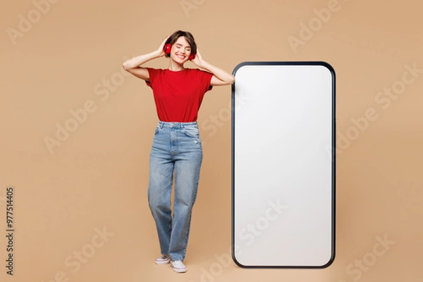 Fototapeta Full body young woman wear red t-shirt casual clothes big huge blank screen mobile cell phone smartphone with area listen to music in headphones isolated on plain beige background. Lifestyle concept.