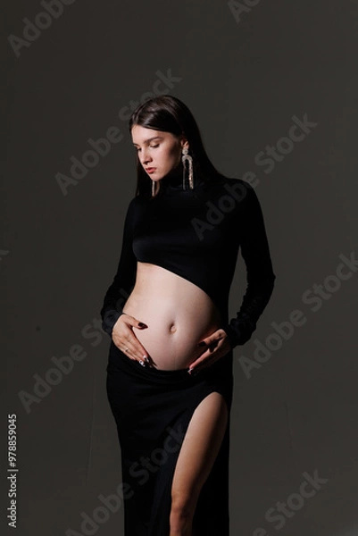Fototapeta Beautiful young pregnant brunette girl. A woman waiting for a baby in a black dress,  in the studio. Dress with a hole in the stomach, pretty pregnant belly.