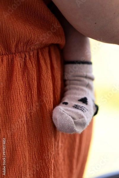 Fototapeta A portrait of the cute leg and foot of a baby wearing a cosy sock pressed safely against the caring mother who is carying the small child.