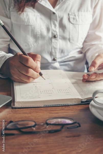 Fototapeta Male hand planner writing daily appointment On Calendar book, working space at home. Man plan and noted schedule (holiday trip) on diary and using smartphone at office desk. Planner concept-vertical
