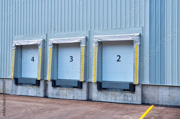 Obraz Side view of a building with three industrial loading docks. Each dock has a closed blue roll-up door and is equipped with an extendable dock leveler at the base.