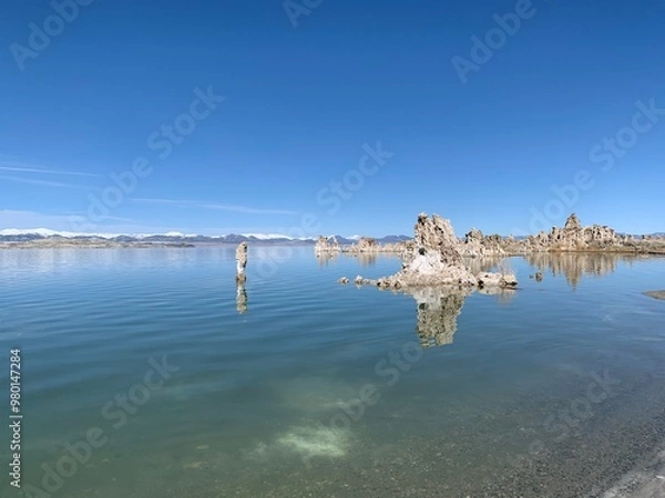 Obraz A wide shot of the calm water surface of Mono Lake on a clear day, revealing the slightly visible lakebed near the shoreline. In the background, snow-capped mountains stand tall on the horizon.