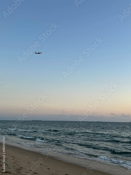 Obraz A serene beach scene with a commercial airplane soaring overhead, captured at Venice Beach, Los Angeles, with the vast Pacific Ocean stretching out to the horizon. The tranquil atmosphere.