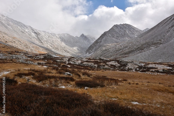 Fototapeta Gentle rocky slopes of the mountain descending into a valley overgrown with red lichens and sprinkled with the first snow on a cloudy autumn morning.