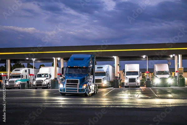 Obraz Commercial big rig semi trucks with semi trailers fill up their fuel tanks at truck stop gas station at night time to continue driving on their routes in the morning