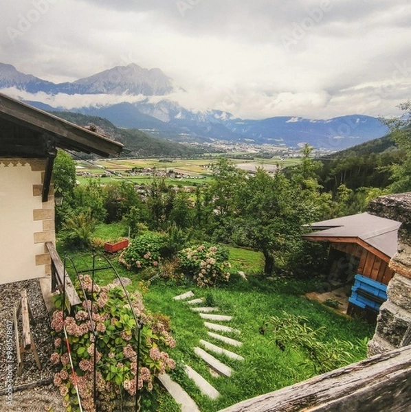 Fototapeta Vibrant, rustic view of an Alpine house in Rietz, Austria, overlooking lush green fields and mountains under a cloudy sky with vintage texture that adds an authentic, old feel to the scene.