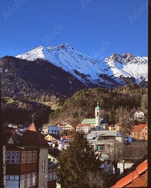 Fototapeta A picturesque view from a window in Innsbruck, Austria, featuring the snow-capped Nordkette Mountain Range towering over the charming alpine town, with a church and colorful houses nestled below.