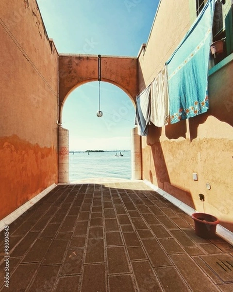Fototapeta A view of the Cannaregio district in Venice, with an arched passage leading to a serene waterfront. Laundry hangs drying in the sunlight, adding a charming touch to this hidden spot.