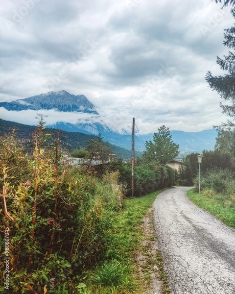 Fototapeta A scenic view of snow-capped mountains in the Alps, captured in Rietz, Austria. The winding path leads through Tirol's lush forests under cloudy skies, highlighting the serene, natural beauty.
