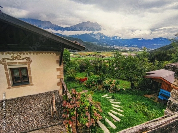 Fototapeta Vibrant, rustic view of an Alpine house in Rietz, Austria, overlooking lush green fields and mountains under a cloudy sky with vintage texture that adds an authentic, old feel to the scene.
