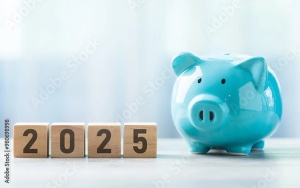 Fototapeta 2025 is written on a wooden block beside a A blue piggy bank on a wooden table against a bright white background. Money planning and Financial planning, savings, and wise money management.