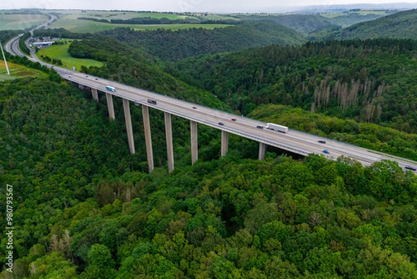 Fototapeta many cargo trucks and cars on the highway on the bridge. asphalt road among green forest and beautiful rain clouds. cargo delivery and transportation concept