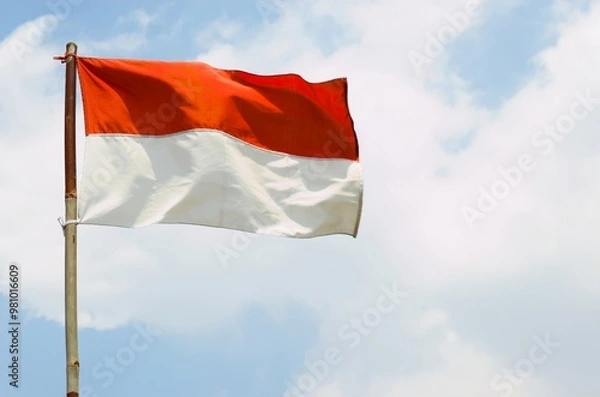 Obraz The red and white Indonesian flag on a bamboo pole flutters in the wind against a backdrop of cloudy blue sky.