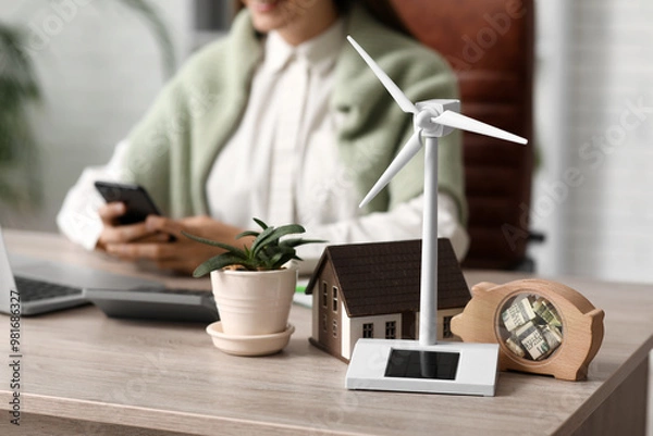Fototapeta Wind turbine with house model, piggy bank and plant on table of female engineer in office, closeup