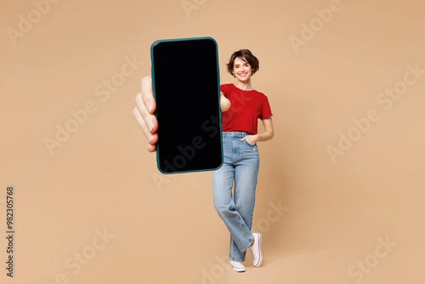 Fototapeta Full body young happy woman she wear red t-shirt casual clothes hold in hand use mobile cell phone with blank screen workspace area isolated on plain pastel light beige background. Lifestyle concept.