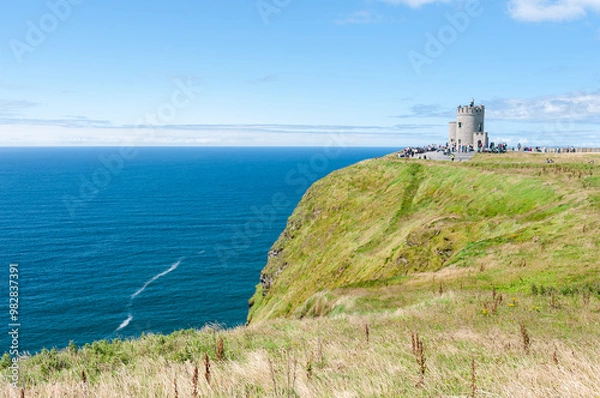 Obraz The Cliffs of Moher stretch along the Atlantic coastline, with a historic  O'Brien's Tower visible at the top. Vertical rock stack rises from the ocean, surrounded by waves, under a cloudy sky.