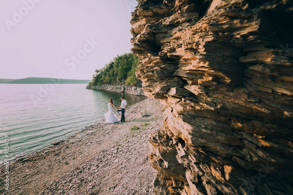 Fototapeta beautiful gorgeous blonde bride and stylish groom holding hands, on the background of a sea
