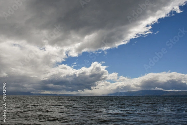 Fototapeta A sky full of beautiful clouds, lit by the sun. Before that it was raining. The rippling waters of the lake overflow in the light. Mountains in the distance.