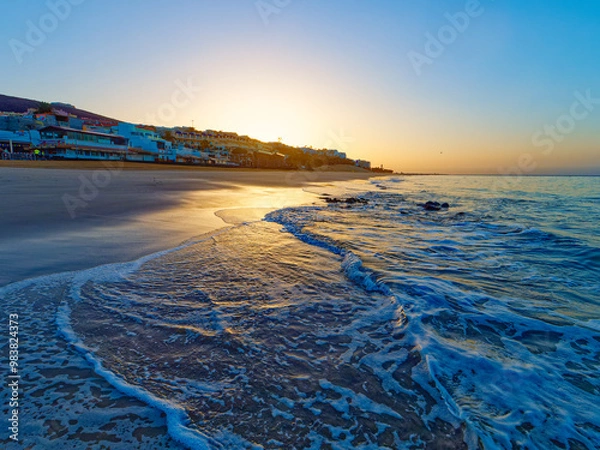 Fototapeta A beautiful sunrise over a sandy beach in Morro Jable on island Fourteventura. The sun is rising over a calm ocean, and the sky is a clear blue. There are palm trees and buildings in the distance.
