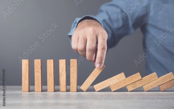 Obraz Businessman uses his hand stopping wooden blocks from falling, representing the domino effect and risk management. Symbolizes taking action to prevent cascading problems and managing risks effectively