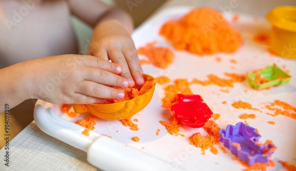 Fototapeta Little child playing with orange kinetic sand at white wooden table, selective focus. Smiling happy child. Hands close up.