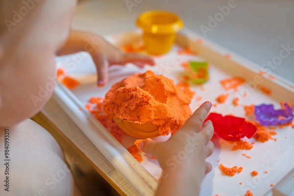 Fototapeta Little child playing with orange kinetic sand at white wooden table, selective focus. Smiling happy child. Hands close up.