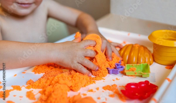 Fototapeta Little child playing with orange kinetic sand at white wooden table, selective focus. Smiling happy child. Hands close up.