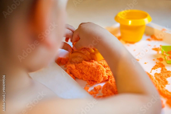 Fototapeta Little child playing with orange kinetic sand at white wooden table, selective focus. Smiling happy child. Hands close up.