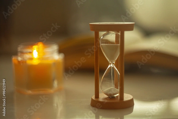 Fototapeta Time to relax - an hourglass in a cozy setting with an open book, and a burning candle, symbolizing taking time to relax and unwind