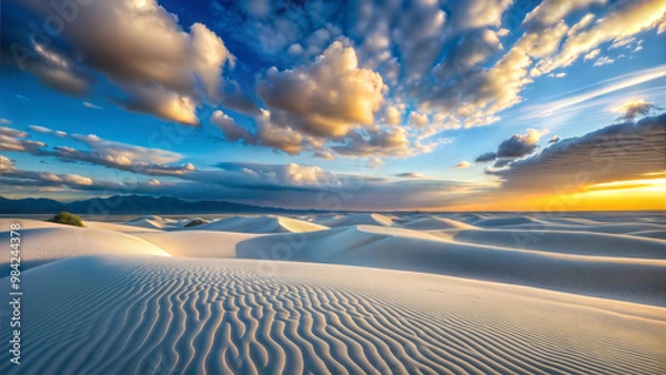 Fototapeta Pristine white sand dunes stretch across the landscape under a dramatic sky. Ripples pattern the sand, while golden sunlight illuminates billowing clouds, creating a surreal and tranquil desert scene.