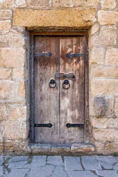 Obraz A weathered wooden door with iron hardware stands firmly within a crumbling stone wall, reflecting the charm of a historic area. The rustic features tell a tale of old-world craftsmanship