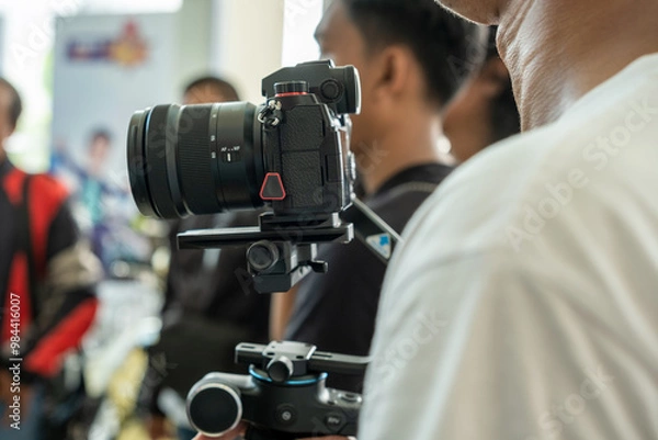 Fototapeta A professional camera mounted on a stabilizer is being set up during a videography or photography session. People are visible in the background, focused on the equipment and preparation.