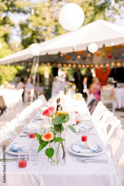 Obraz A backyard set up for a wedding reception with a tent in the background, and the table is set up with white tablecloths, flowers, and porcelain plates. 