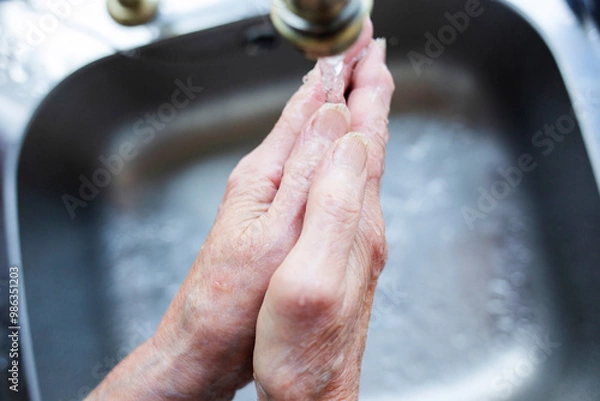 Obraz Close-up of elderly lady washing her hands under a running tap against a silver shade sink 